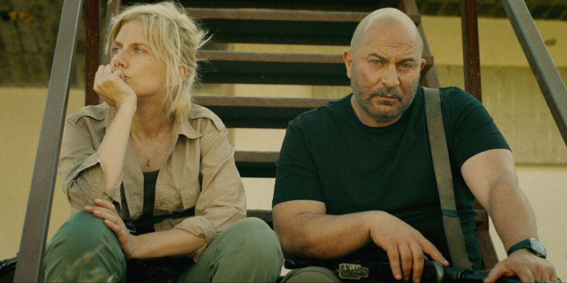 "Two serious people sit on outdoor metal stairs in casual, tactical clothing, appearing tired or contemplative, with sunlight casting shadows behind them against a plain wall in a rugged environment."