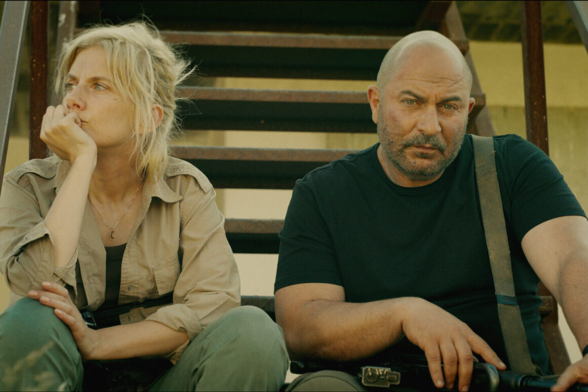 "Two serious people sit on outdoor metal stairs in casual, tactical clothing, appearing tired or contemplative, with sunlight casting shadows behind them against a plain wall in a rugged environment."