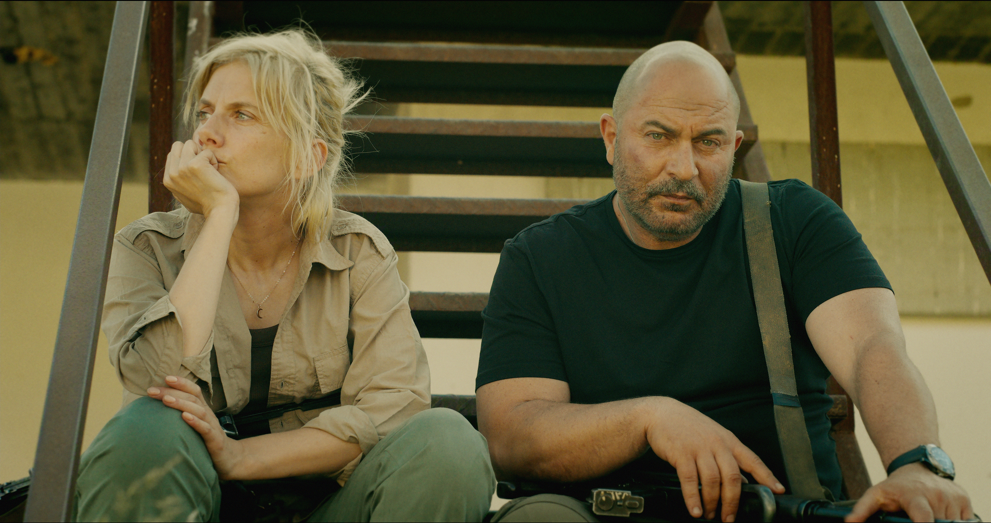 "Two serious people sit on outdoor metal stairs in casual, tactical clothing, appearing tired or contemplative, with sunlight casting shadows behind them against a plain wall in a rugged environment."