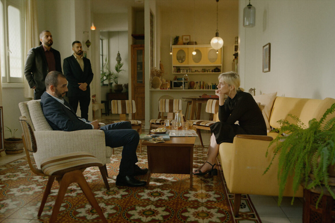 "Four people in a living room with mid-century decor; two seated and talking, two standing behind, surrounded by vintage furniture, patterned floor, plants, and warm lighting."